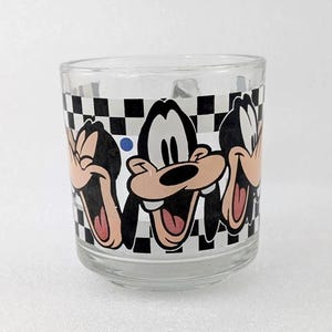 May include: A clear glass mug featuring a repeating graphic print of the cartoon character Goofy in various laughing poses. The design is set against a black and white checkered background with a small blue dot accenting the pattern.