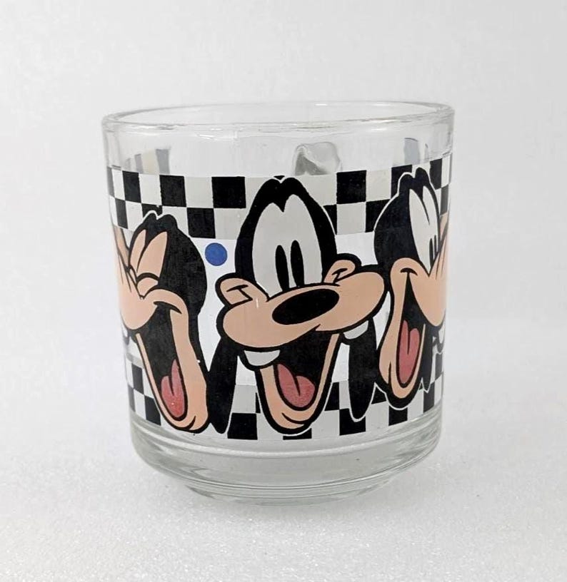 May include: A clear glass mug featuring a repeating graphic print of the cartoon character Goofy in various laughing poses. The design is set against a black and white checkered background with a small blue dot accenting the pattern.