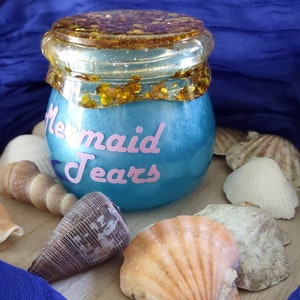 May include: A blue jar with pink text reading "Mermaid Tears", topped with a gold glitter lid. Seashells surround the jar against a blue fabric backdrop.