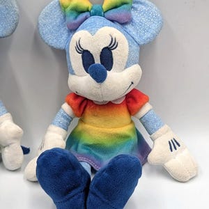 May include: A plush Minnie Mouse doll with a rainbow bow, dress, and blue ears and shoes. The doll has white gloves and a white face with black eyelashes and a blue nose. The doll is sitting down.