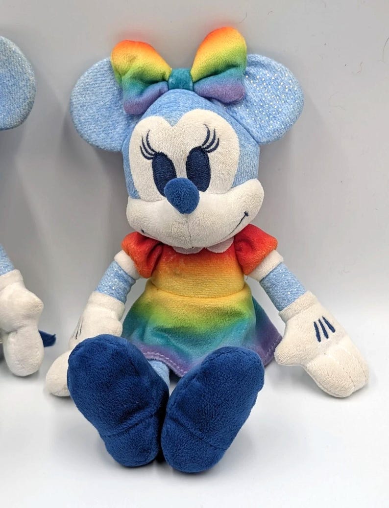 May include: A plush Minnie Mouse doll with a rainbow bow, dress, and blue ears and shoes. The doll has white gloves and a white face with black eyelashes and a blue nose. The doll is sitting down.