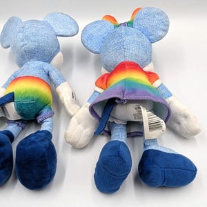 May include: Two plush toys of a mouse character, viewed from the back. The toys are primarily light blue with white accents. One toy has a rainbow-colored skirt and the other has rainbow-colored shorts. Both have large blue feet.