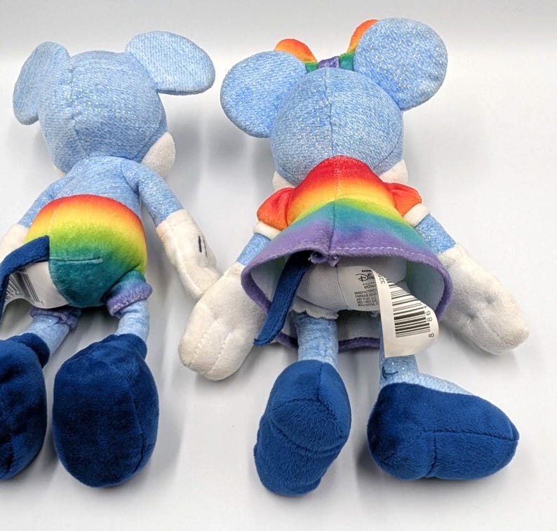 May include: Two plush toys of a mouse character, viewed from the back. The toys are primarily light blue with white accents. One toy has a rainbow-colored skirt and the other has rainbow-colored shorts. Both have large blue feet.