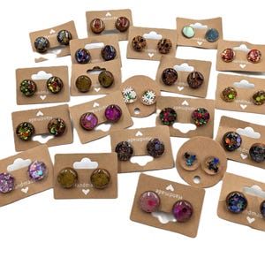 May include: A collection of handmade stud earrings in various colors and designs. The earrings are displayed on small, brown cardboard earring cards. The earrings feature glitter, floral, and abstract patterns.