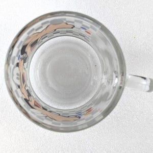 May include: A clear glass mug viewed from directly above, featuring a repeating checkered pattern around the exterior rim. The design includes partial illustrations of a cartoon character with tan skin tones and small blue and red circular accents.