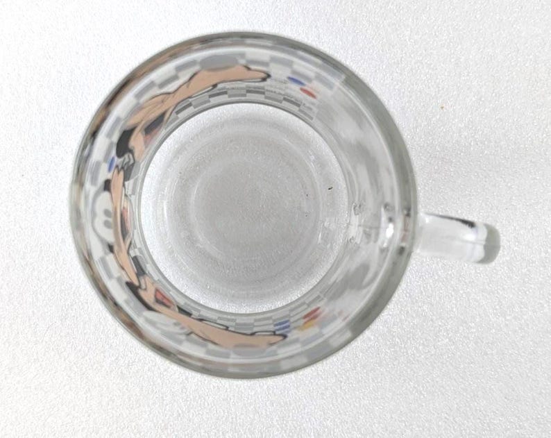 May include: A clear glass mug viewed from directly above, featuring a repeating checkered pattern around the exterior rim. The design includes partial illustrations of a cartoon character with tan skin tones and small blue and red circular accents.