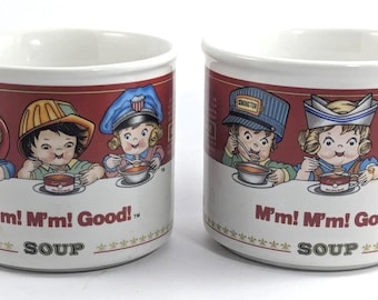 May include: Two white ceramic mugs featuring a red band with illustrations of children wearing various hats and eating soup. The text M'm! M'm! Good! and SOUP is printed on the front of each mug. The mugs have a handle on the side.