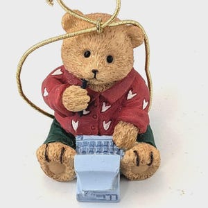 Op de afbeelding: Een bruin teddybeerornament met een rood shirt met witte hartjes en een groene broek. De beer houdt een klein zwart voorwerp vast en zit voor een lichtblauw voorwerp. Een gouden touwtje is bevestigd om op te hangen.