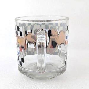 May include: A clear glass mug featuring a wrap-around graphic of a cartoon character with tan skin and black ears. The design includes a black and white checkered border along the top and bottom, accented by small red, blue, and yellow circular dots.