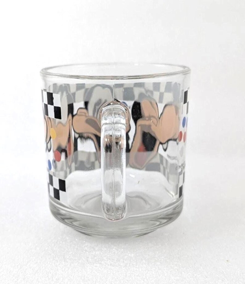 May include: A clear glass mug featuring a wrap-around graphic of a cartoon character with tan skin and black ears. The design includes a black and white checkered border along the top and bottom, accented by small red, blue, and yellow circular dots.