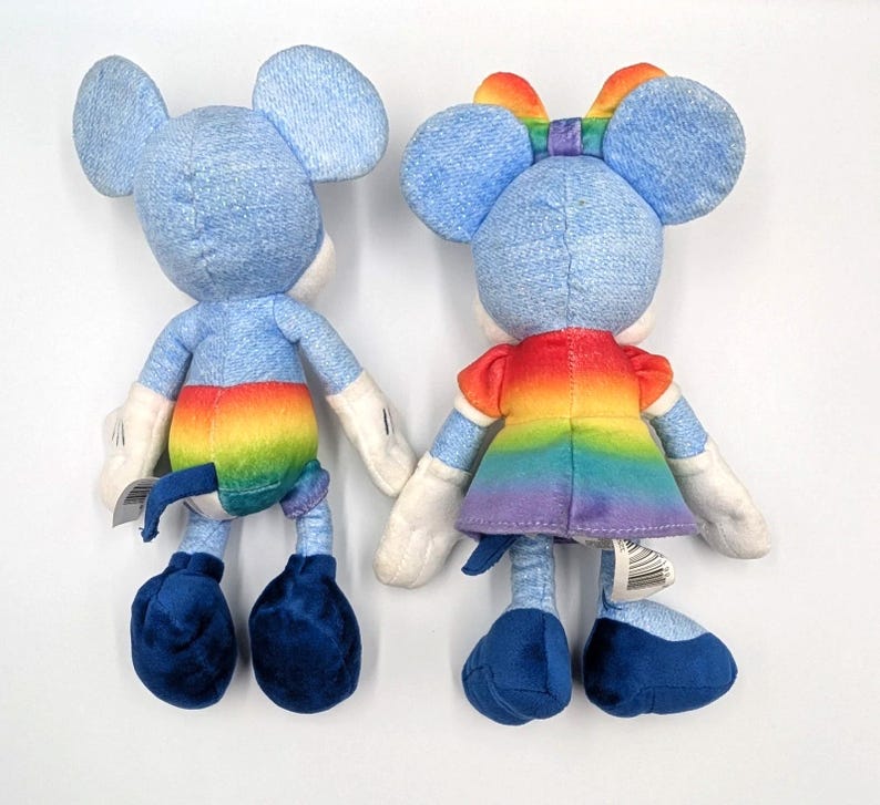 May include: Two plush toys of a mouse character, one with a rainbow-colored tail and the other with a rainbow dress and bow. Both toys have blue denim-like fabric for their bodies, large ears, and white gloves.