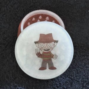 May include: A round, two-piece grinder with a cartoon image of Freddy Krueger on the lid. The lid is white with a light blue border. The grinder's base is brown with a textured surface.