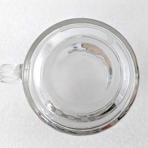 May include: A clear glass mug viewed from directly above, featuring a circular base and a side handle. The bottom of the mug displays a subtle, embossed grid pattern around the perimeter, with a smooth, transparent center section.