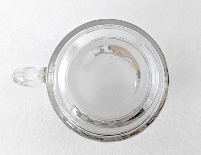 May include: A clear glass mug viewed from directly above, featuring a circular base and a side handle. The bottom of the mug displays a subtle, embossed grid pattern around the perimeter, with a smooth, transparent center section.