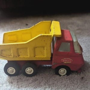 May include: A vintage red and yellow Tonka dump truck toy. The truck features a yellow dump bed and a red cab with the Tonka logo. The toy has black tires with white rims and shows signs of wear.
