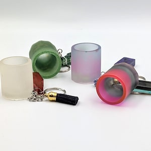 May include: A collection of four cylindrical keychains featuring translucent and opaque finishes in frosted white, textured green, gradient pink and purple, and a bold pink and black design, each attached to a metal ring with a black tassel.