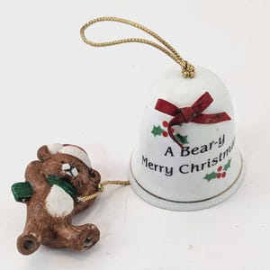 May include: A white ceramic Christmas bell with a gold string and a red bow. The bell has the text "A Bear-y Merry Christmas" and holly accents. A brown bear figurine with a Santa hat and green scarf is attached.