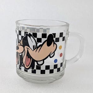 May include: A clear glass mug featuring a wrap-around graphic of the cartoon character Goofy. The design includes a black and white checkered border along the top and bottom, with small colored dots in yellow, red, and blue on the side.