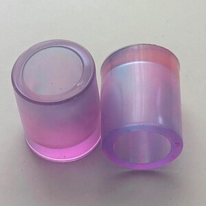 May include: Two cylindrical resin objects with a smooth, translucent finish. The items feature a gradient colour transition from a soft pink at the base to a pale blue and light purple hue at the top, with a hollow centre and thick, rounded walls.