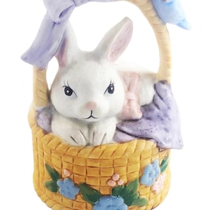 May include: A ceramic Easter basket with a white bunny inside. The basket is yellow with blue flowers and green leaves. A light blue bird sits on the handle. A light purple bow is on top of the handle.