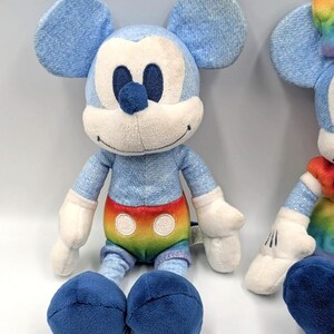 May include: Two plush Mickey Mouse toys. The toys have blue denim-like fabric for the head, ears, arms, and legs. The belly features a rainbow design. The face is white with black eyes and a blue nose. The feet are dark blue.