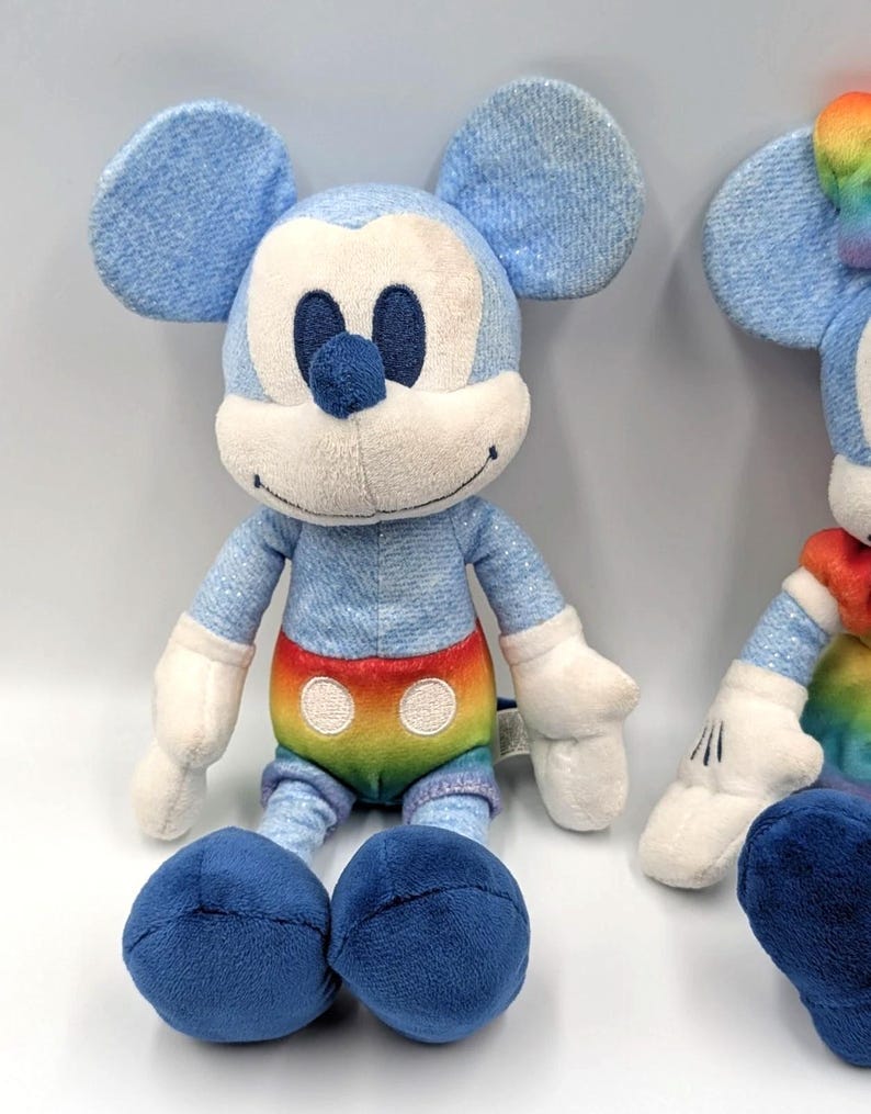 May include: Two plush Mickey Mouse toys. The toys have blue denim-like fabric for the head, ears, arms, and legs. The belly features a rainbow design. The face is white with black eyes and a blue nose. The feet are dark blue.
