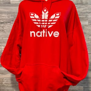 Könnte beinhalten: Ein leuchtend roter Kapuzenpullover mit einer weißen Grafik eines stilisierten Dreiblatt-Logos über dem Wort native in einer fetten serifenlosen Schrift. Das Kleidungsstück verfügt über eine Kängurutasche, lange Ärmel und rote Kordeln.