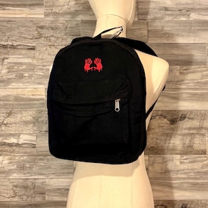 May include: Black backpack with a front pocket and a zipper closure. The backpack features a red embroidered design of two hands. The bag is displayed on a mannequin against a gray and brown wood-look background.