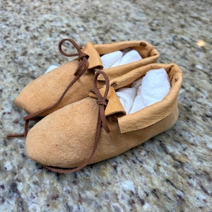 May include: A pair of tan suede moccasins featuring a soft, flexible construction with a folded collar and thin brown leather laces tied in a bow at the front. The interior of each shoe is stuffed with white tissue paper to maintain its shape.
