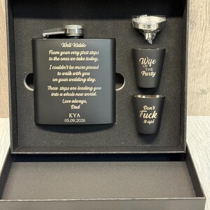 May include: A black matte gift set containing a metal flask, two shot glasses, and a funnel. The flask is engraved with the text: Well Kiddo, From your very first steps to the ones we take today, I couldn't be more proud to walk with you on your wedding day. These steps are leading you into a whole new world. Love always, Dad, KYA, 05.09.2026. The shot glasses feature the text: Wife OF THE Party and Don't Fuck it up!