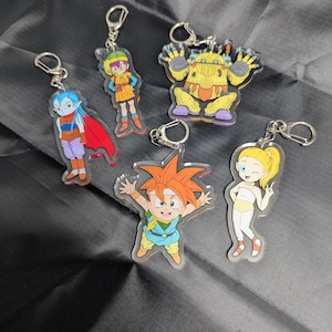 May include: Five colorful anime character keychains with silver key rings. The characters are a girl with orange hair, a boy with blue hair, a boy with orange hair, a girl with blonde hair, and a robot.
