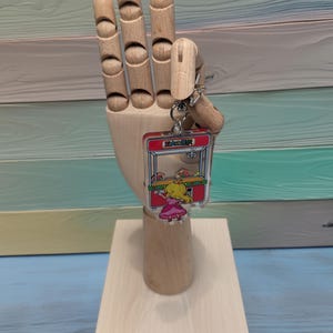 May include: A clear acrylic keychain featuring a cartoon character playing a claw machine game. The keychain is red, yellow, and green with the text "お菓子掴み" in Japanese.