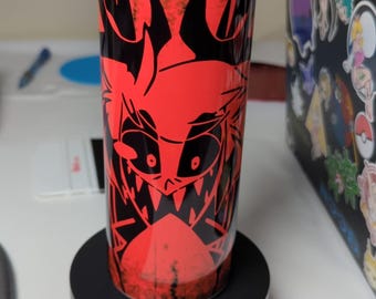 May include: A red and black stainless steel tumbler with a graphic of a cartoon character with horns and a wide grin. The tumbler has a black lid and is sitting on a black coaster.