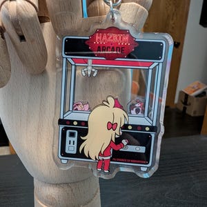 May include: Clear acrylic keychain featuring a cartoon arcade claw machine with the text "HAZBIN ARCADE". The keychain has a silver metal ring and a cartoon character in a red outfit. The arcade machine has a red and black design.