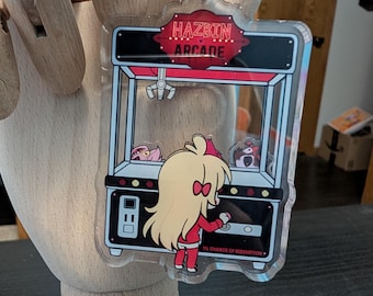 Puede incluir: Llavero acrílico transparente con un diseño de máquina de garras de arcade de dibujos animados con el texto "HAZBIN ARCADE". El llavero tiene un anillo de metal plateado y un personaje de dibujos animados con un atuendo rojo. La máquina recreativa tiene un diseño rojo y negro.