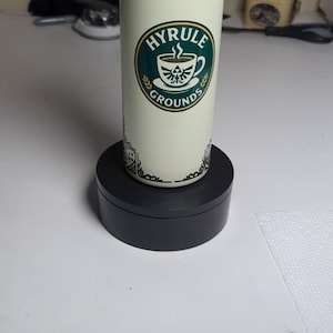 May include: A cylindrical tumbler with a cream-colored background and the words "HYRULE GROUNDS" in a green and gold circular logo. The tumbler is decorated with black ornate designs and sits on a black rotating base. The tumbler is designed for beverages.