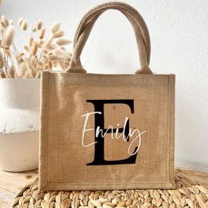 May include: A brown burlap tote bag with a black letter "E" and the name "Emily" printed in white.