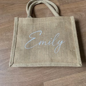 Pode incluir: Uma bolsa de compras de juta marrom natural com al&ccedil;as brancas e o nome "Emily" impresso em escrita cursiva branca.