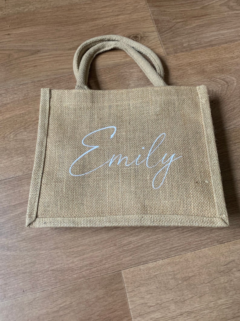 Pode incluir: Uma bolsa de compras de juta marrom natural com al&ccedil;as brancas e o nome "Emily" impresso em escrita cursiva branca.