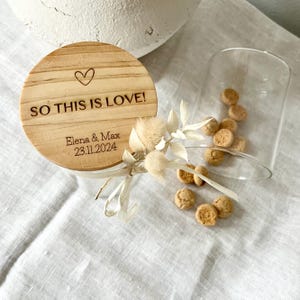 May include: A wooden lid with the text "So this is love!" and the names "Elena & Max" and the date "23.11.2024" engraved on it. The lid is sitting on top of a glass jar.