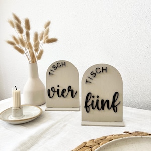May include: Two wooden table number signs with arched tops. The signs are cream-colored with black lettering that reads "TISCH" above and "vier" and "fünf" below. A vase with dried flowers and a candle are also in the image.