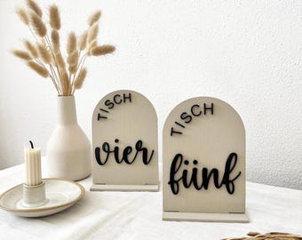 Pode incluir: Dois sinais de número de mesa de madeira creme com topos arqueados. Os sinais têm letras pretas que dizem "TISCH" acima e "vier" e "fünf" abaixo. Um vaso com flores secas e uma vela também estão na imagem.