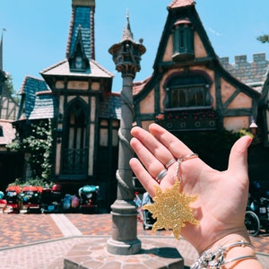 May include: A gold glitter sun keyring with a black string. The keyring is held in a hand in front of a building with a tower.