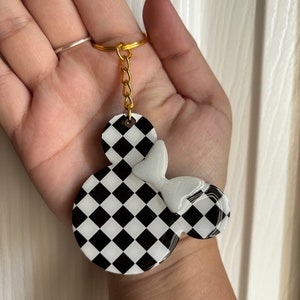 May include: Checkerboard patterned black and white resin keychain with a white bow. The keychain is shaped like a cartoon mouse head.