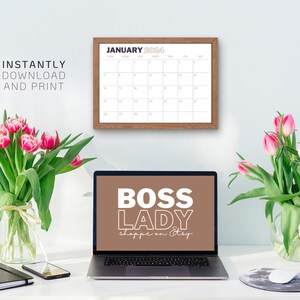 Op de afbeelding: Een laptop met een bruine achtergrond op het scherm waarop "BOSS LADY shoppe on Etsy" staat. De laptop staat op een wit bureau met een kalender, twee vazen met roze tulpen, een zwart notitieboekje en een mobiele telefoon.