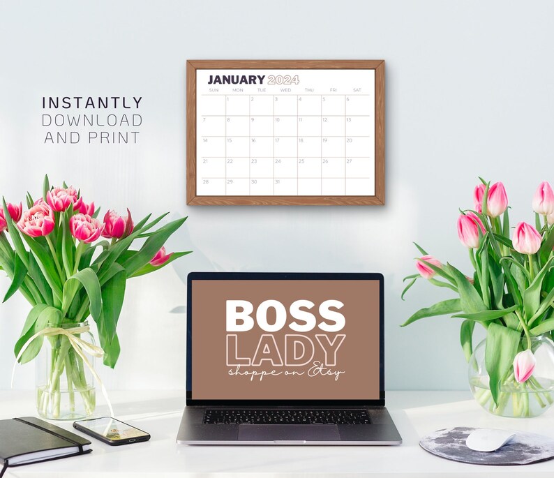 Op de afbeelding: Een laptop met een bruine achtergrond op het scherm waarop "BOSS LADY shoppe on Etsy" staat. De laptop staat op een wit bureau met een kalender, twee vazen met roze tulpen, een zwart notitieboekje en een mobiele telefoon.