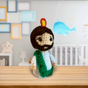 May include: Crocheted amigurumi doll of a religious figure with a green robe, brown hair, and a beard. The doll is wearing a red and yellow halo.