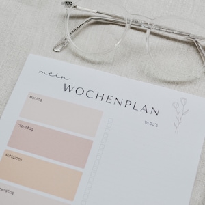 Könnte beinhalten: Ein Wochenplaner mit den Worten "mein WOCHENPLAN" in Schwarz gedruckt. Der Planer hat Abschnitte für jeden Wochentag, mit einer floralen Illustration und Platz für To-Do-Listen. Eine klare Brille liegt obenauf.