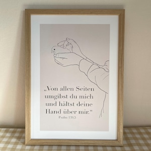 Könnte beinhalten: Ein gerahmter Kunstdruck mit einer Strichzeichnung von zwei Händen, die einen kleinen Vogel halten. Unter der Illustration steht der deutsche Text: „Von allen Seiten umgibst du mich und hältst deine Hand über mir.“ Psalm 139,5. Der Rahmen ist aus hellem Holz.