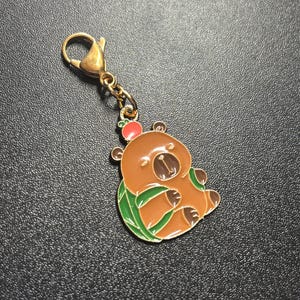 May include: A brown bear charm with a gold-colored clasp and chain. The bear has a red apple on its head and is surrounded by green leaves. The charm is made of metal and has a glossy finish.